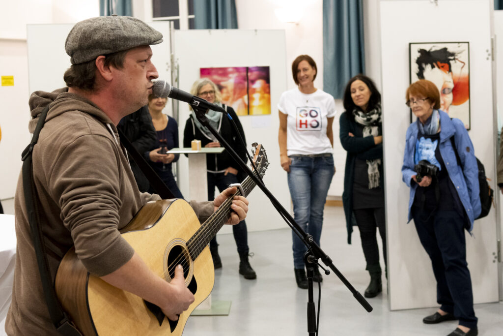 Ein Gitarrist steht am Mikrofon bei einer Eröffnungsfeier der Gostenhofer Ateliertage, im Hintergrund stehen Menschen und schauen ihm zu